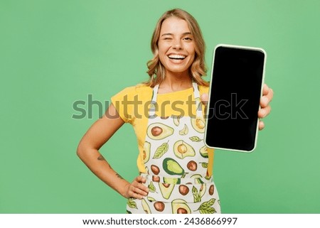 Young smiling housewife housekeeper chef cook baker woman wear apron yellow t-shirt hold use blank screen mobile cell phone blink eye isolated on plain pastel green background. Cooking food concept