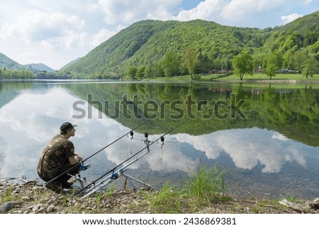 Fishing adventures, carp fishing on lake. Man crouching next to fishing rods waiting to catch a fish. Rod pod with two fishing rods and electronic bite alarms. Selective focus on the fisherman
