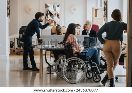 A cross generational group of professionals engage in a business meeting. The image includes a person using a wheelchair, depicting inclusivity and teamwork in a modern office setting. Royalty-Free Stock Photo #2436878097
