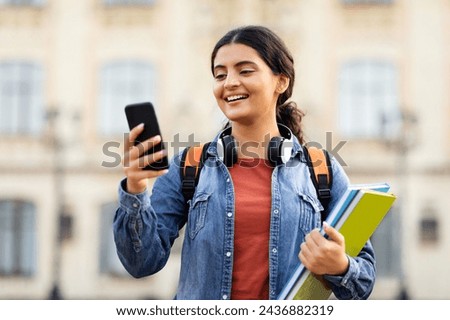 Online learning, e-education concept. Happy young indian lady student using phone outdoors, carrying backpack and textbooks, going to university, checking her schedule online Royalty-Free Stock Photo #2436882319