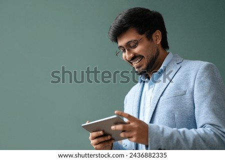 Happy hindu businessman smiles websurfing on digital tablet computer, standing against dark green wall in office, browsing business application, closeup. Empty space for online offers Royalty-Free Stock Photo #2436882335