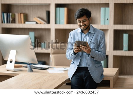 Cheerful Indian professional businessman websurfing on digital tablet during work in contemporary office environment, browsing web at modern workplace. Internet and technology for business Royalty-Free Stock Photo #2436882457
