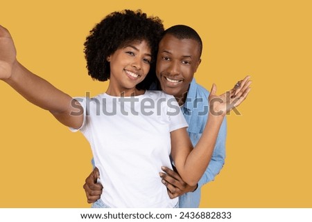 A radiant young couple capturing a cheerful selfie, with the woman stretching out her arm to take the photo and the man gently holding her, against a mustard yellow background