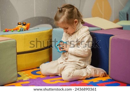 Little girl sits on colorful play mat, fully absorbed in solving handheld puzzle toy in preschool environment. Royalty-Free Stock Photo #2436890361