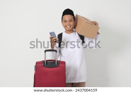 Portrait of excited Asian muslim man carrying cardboard box and suitcase while holding smartphone. Going home for Eid Mubarak. Isolated image on white background