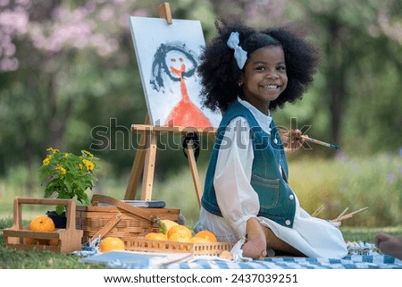 African little girl holds brush and paints picture on easel at park, very happy and smiling at camera, her younger brother sat next to her.