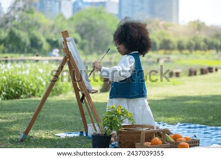 African little girl holds brush and paints picture on easel at park, very happy and smiling at her work, spend the holidays picnicking and painting