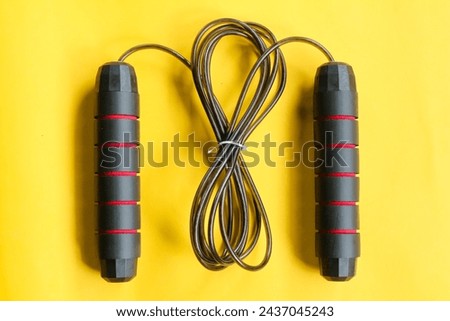 The top view of a black skipping rope lies down on the yellow background. Jump. Health. Sport. Healthy. Background. Activity. Equipment. Play. Skip. Athlete. Strength. Workout. Cable

