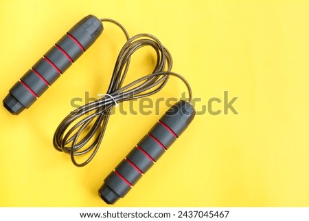 The top view of a black skipping rope lies down on the yellow background. Jump. Health. Sport. Healthy. Background. Activity. Equipment. Play. Skip. Athlete. Strength. Workout. Cable


