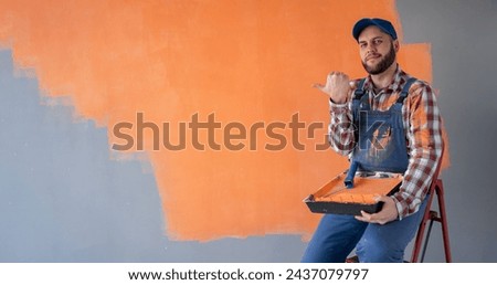 Positive professional male painter with roller pointing at blank space on orange wall sitting on ladder and looking at camera. Banner. Copy space