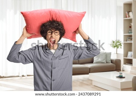 Angry young man in pajamas holding a pillow over head and screaming at home