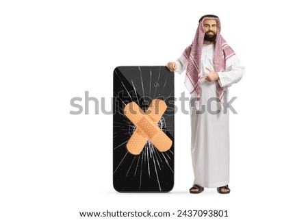 Saudi arab man in ethnic clothes pointing at a cracked screen on a smartphone fixed with a bandage isolated on white background
