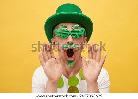 Shocked scared man wearing green hat and clover glasses standing isolated over yellow background saying OMG looking with open mouth and frighten face raising hands