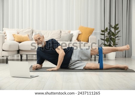 Senior man doing exercise with fitness elastic band near laptop on mat at home Royalty-Free Stock Photo #2437111265