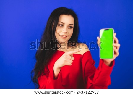 Excited young woman in red top hold and show blank phone green screen with copy space for app, isolated on blue background. Happy amazed woman with wow emotions, recommend app or some product.