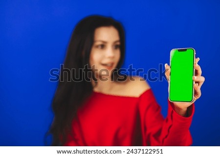 Excited young woman in red top hold and show blank phone green screen with copy space for app, isolated on blue background. Happy woman with wow emotions, recommend app or some product. Focus on phone