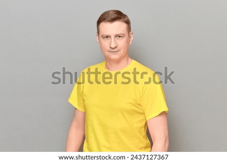 Studio portrait of blond mature man wearing yellow T-shirt, with happy expression on face, with positive facial emotion, smiling cheerfully, holding hands along body, standing over gray background