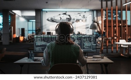 Creative Office: Professional Female Programmer Uses Headphones, Working on Desktop Computer. Focused Coder, Software Engineer Developing App, Video Game. Listening to Podcast or Music. Back View. Royalty-Free Stock Photo #2437129381