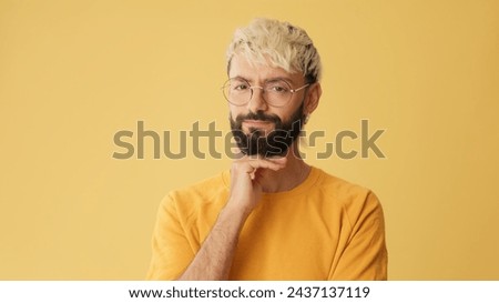 Guy with glasses, dressed in yellow T-shirt, listens carefully and disagree, isolated on yellow background in studio