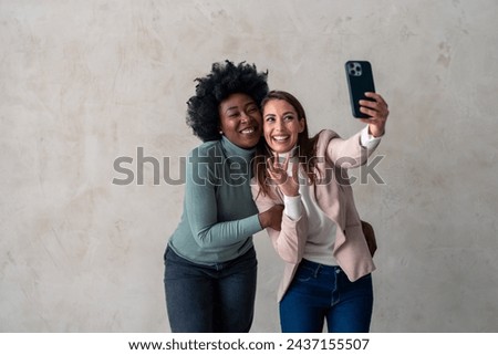 Ecstatic vibrant young women having video call on mobile phone, embracing, waving hand while standing together against a studio background.