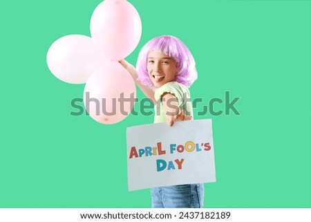Funny little girl in wig with balloons and card showing tongue on green background. April Fools' Day celebration Royalty-Free Stock Photo #2437182189