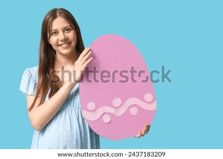 Beautiful young pregnant woman with paper Easter egg on blue background