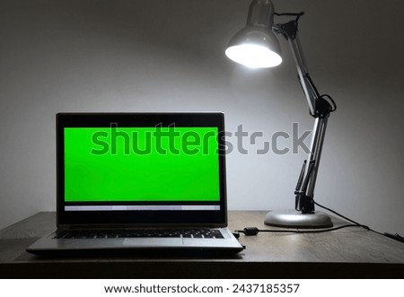 mock up. Laptop with blank green screen, and office on wooden desk. Blank computer monitor on wood table with lamp light. isolated on white wall background. mockup. front view. night evening scene.