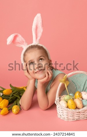 Portrait of cute smiling girl with Bunny ears and yellow Easter eggs, near yellow tulips, isolated on pink background. Happy Easter
