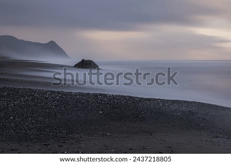 A rocky beach with a large rock in the foreground. The sky is cloudy and the water is calm