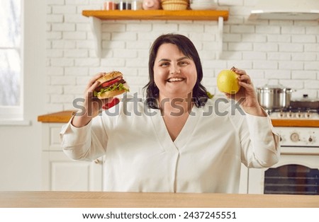Young overweight plus size fat woman sitting in the kitchen at home holding a burger in one hand and an apple in other thinking what to choose. Diet restrictions, losing weight concept. Royalty-Free Stock Photo #2437245551
