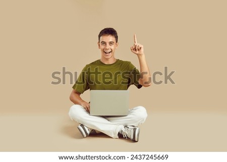 Portrait happy clever adolescent high school student boy with laptop computer sitting on floor on beige studio background, looking at camera, smiling, pointing up, saying Aha eureka I have genius idea Royalty-Free Stock Photo #2437245669