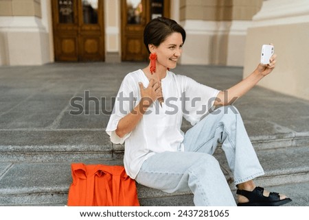 attractive woman walking in street in summer fashion trend style outfit smiling happy using smartphone taking pictures on camera, white blouse, orange bag, purse, earrings, Europe vacation