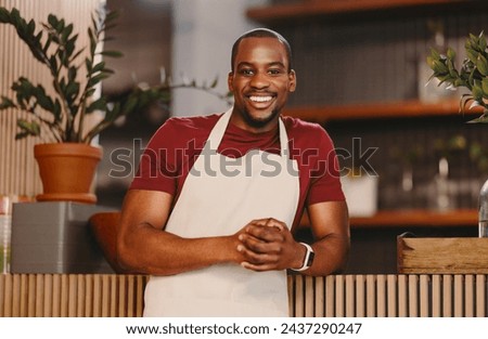 Young man looking at the camera with a bright smile while wearing a white apron in a cozy cafe. Male entrepreneur embodying success, hospitality, and small business owner vibes. Royalty-Free Stock Photo #2437290247