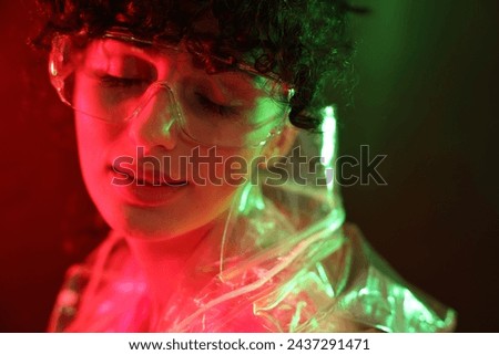 Beautiful young woman in transparent coat and sunglasses posing on color background in neon lights, closeup