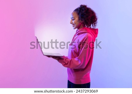 Cheerful young lady holding an open laptop with glowing screen, standing against vibrant pink and blue gradient background, looking content and tech-savvy Royalty-Free Stock Photo #2437293069