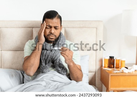 Unhealthy young african american guy with warm scarf around his neck sitting in bed, checking body temperature with electronic thermometer, touching head, suffering from fever, copy space. Cold flu Royalty-Free Stock Photo #2437293093