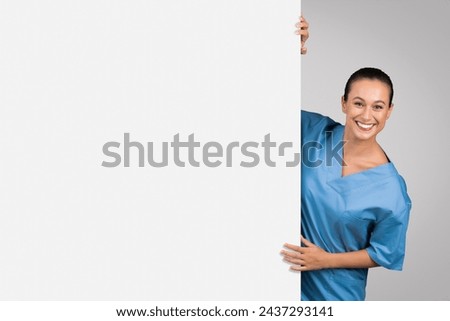 Positive female nurse in blue attire playfully peeks around large blank board, perfect for healthcare information or medical ad on grey background, free space