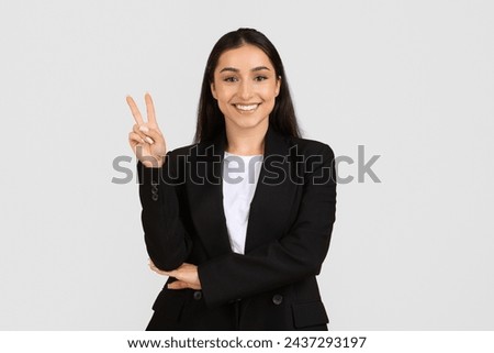 Portrait of professional young woman in business suit smiling confidently while making peace sign against light grey background
