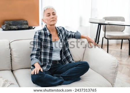 Relaxed Caucasian middle-aged woman having rest on the couch in the living room. Mature housewife sitting in lotus position at home alone. Morning routine Royalty-Free Stock Photo #2437313765