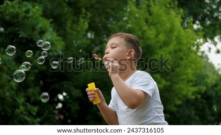 Preschooler boy spends time blowing soap bubbles creating playful atmosphere slow motion. Playing in summer city park cheerful boy happily blows bubbles. Young boy delights in blowing soap bubbles Royalty-Free Stock Photo #2437316105