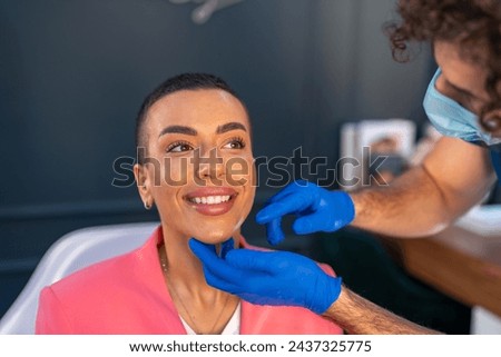 Professional male beautician explaining skin procedures before mesotherapy or skin treatment. Smiling young woman at aesthetic medical clinic. Royalty-Free Stock Photo #2437325775