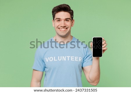 Young man wearing blue t-shirt white title volunteer hold use mobile cell phone with blank screen isolated on plain pastel green background. Voluntary free work assistance help charity grace concept