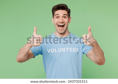Young shocked happy man wear blue t-shirt white title volunteer showing thumb up like gesture isolated on plain pastel light green background. Voluntary free work assistance help charity grace concept