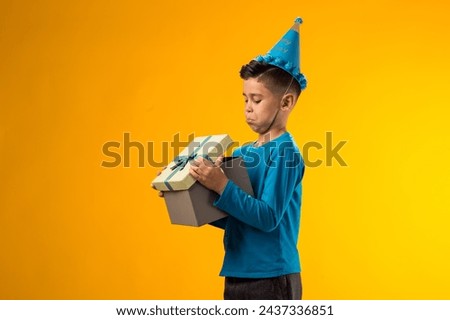 Portrait of child boy with birthday cap on head holding giftbox and looking inside box over yellow background. Birthday and celebration concept