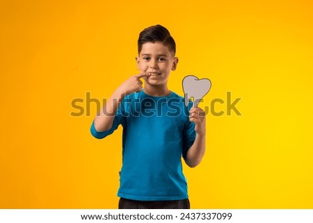 Portrait of child boy holding papercraft tooth and pointing finger at mouth, over yellow background. Dental health concept