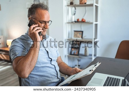 Middle aged Caucasian man talking on the phone while working on laptop at home