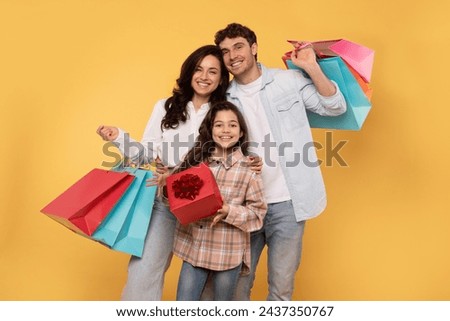 Cheerful family of three people holding bright shopper bags, excited teen girl holding wrapped present box, posing isolated on yellow studio wall