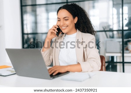 Positive busy brazilian or hispanic curly haired business lady, ceo, sales manager, sits in modern office space with a laptop, looking at the screen, talking by smart phone with client, smile friendly Royalty-Free Stock Photo #2437350921