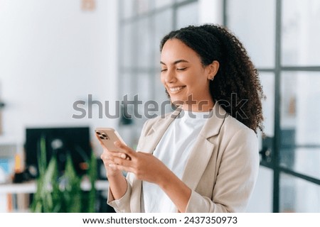 A beautiful positive brazilian or hispanic young woman with curly hair in an elegant jacket, stands in a modern office space, uses her smartphone, messaging with friends on social networks, smiles Royalty-Free Stock Photo #2437350973