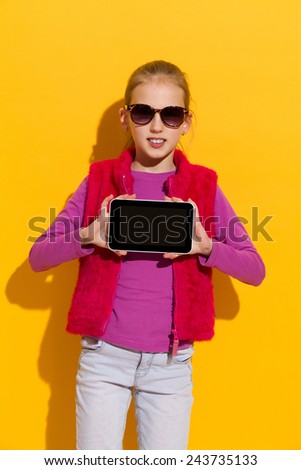 Young blond girl showing a digital tablet. Three quarter length studio shot on yellow background.